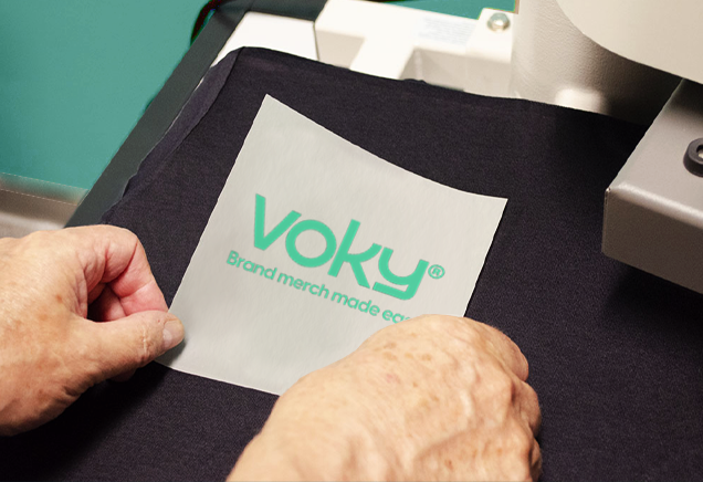 Voky-logotypen tryckt p&aring; pappersh&aring;llare, placerad p&aring; svart tyg f&ouml;r transfertryck.