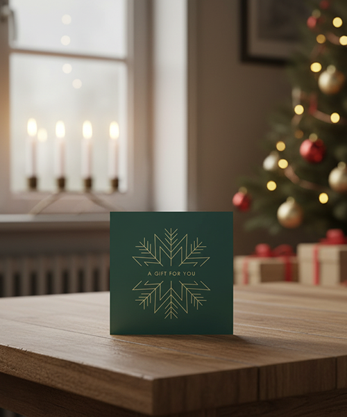 Ett grönt presentkort med gyllene snöflinga och texten "A GIFT FOR YOU" på ett bord, med adventsljusstake och julgran i bakgrunden.