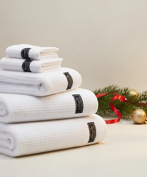 En stapel vita frottéhanddukar med svarta lappar, bredvid en julgranskvist och julgranskulor.